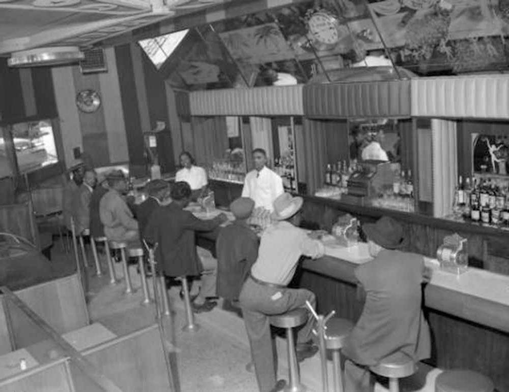 Interior photograph of one male (left) and one female bartending at the 715 Club located at 715 East 26th (twenty-sixth) Avenue in the Five Points neighborhood. A line of male customers fill the seats at the bar. They wear fedora and newsboy hats.